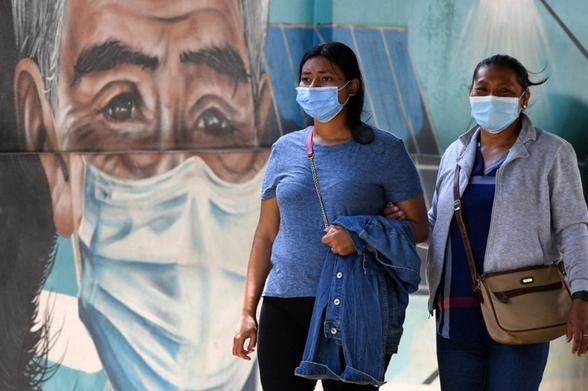 Two women wearing surgical masks walk in front of a mural depicting an older man wearing an surgical mask. Two women wearing surgical masks walk in front of a mural depicting an older man wearing an surgical mask.