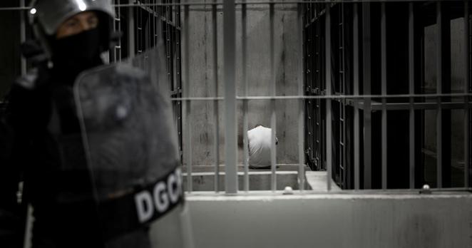 A person sits inside a cell while a guard with a riot shield stands outside