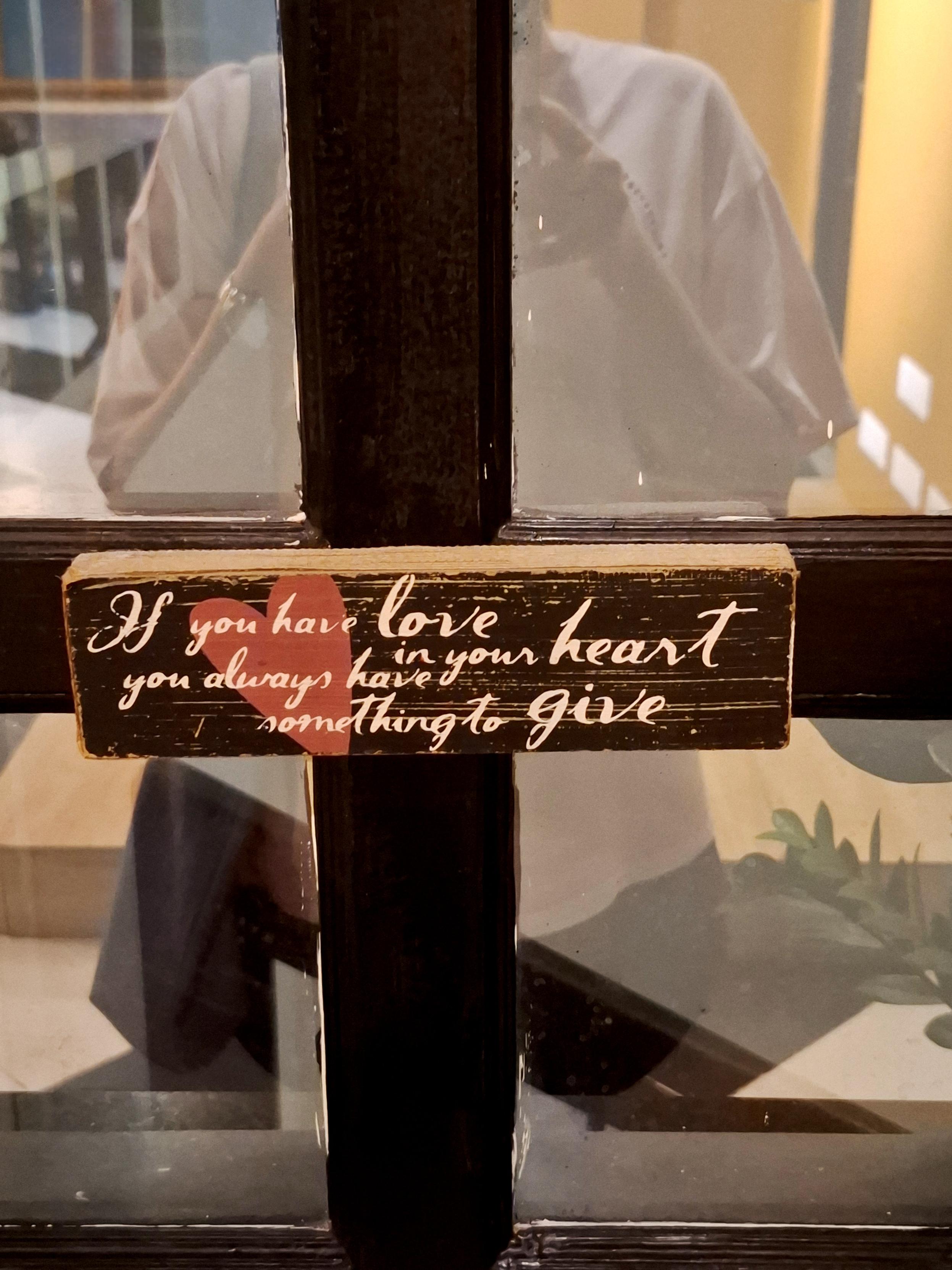 Quote on a block of wood on a glass door which says 'If you have love in your heart, you always have something to give.' My reflection while taking the picture is visible. 
Its such a good quote and its rooted in deep truth. Capitalism makes us believe in the monetary value of things but the best experiences and memories are priceless, they come from the place of love.